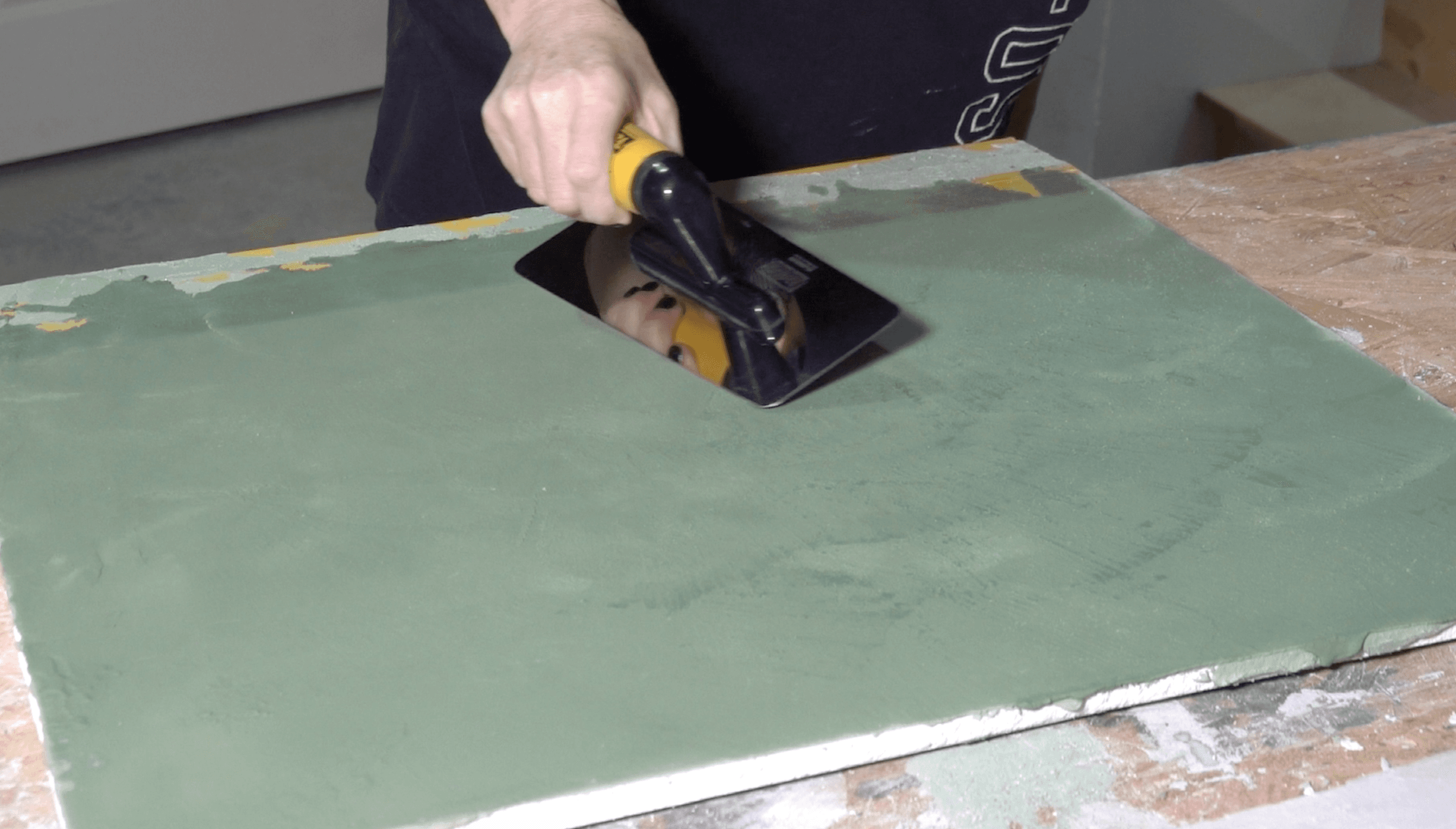 Caroline at the studio bench, drawing a steel trowel across a large sage lime-plaster board.